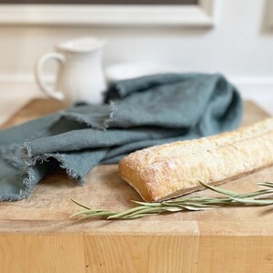 Puede incluir: Una tabla de cortar de madera r&uacute;stica con una hogaza de pan, una rama de vegetaci&oacute;n y un pa&ntilde;o de lino azul doblado.