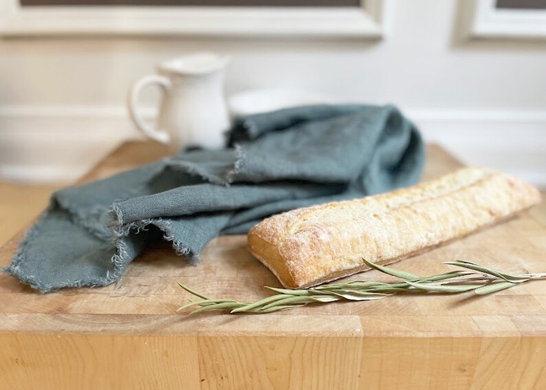 Puede incluir: Una tabla de cortar de madera r&uacute;stica con una hogaza de pan, una rama de vegetaci&oacute;n y un pa&ntilde;o de lino azul doblado.