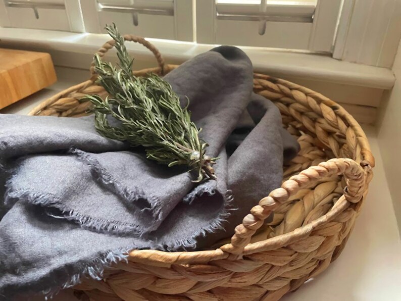 Op de afbeelding: Een geweven rieten mand met twee handvatten, gevuld met een grijze linnen doek en een takje rozemarijn.