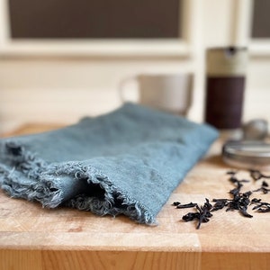 Puede incluir: Un pa&ntilde;o de cocina de lino azul con bordes deshilachados se encuentra sobre una tabla de cortar de madera con hojas de t&eacute; sueltas esparcidas a su alrededor.
