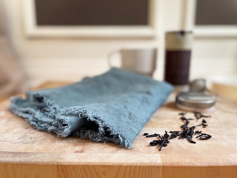 Puede incluir: Un pa&ntilde;o de cocina de lino azul con bordes deshilachados se encuentra sobre una tabla de cortar de madera con hojas de t&eacute; sueltas esparcidas a su alrededor.