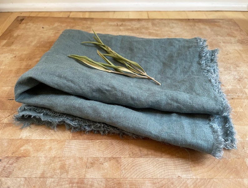 Puede incluir: Un trozo de tela de lino de color turquesa doblado con bordes deshilachados. Una rama de follaje verde descansa sobre la tela.