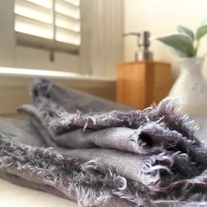 Op de afbeelding: Een grijze linnen keukendoek met rafelige randen. De handdoek is gevouwen en heeft een zachte, textuur.