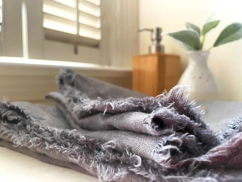 Op de afbeelding: Een grijze linnen keukendoek met rafelige randen. De handdoek is gevouwen en heeft een zachte, textuur.