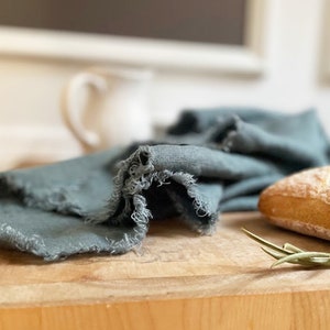 Puede incluir: Una toalla de cocina de lino azul verdoso con bordes deshilachados est&aacute; doblada sobre una tabla de cortar de madera. Tambi&eacute;n hay una hogaza de pan y una rama de verdura en la tabla.