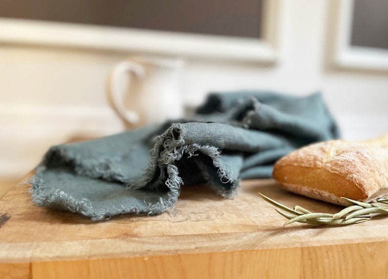 Puede incluir: Una toalla de cocina de lino azul verdoso con bordes deshilachados est&aacute; doblada sobre una tabla de cortar de madera. Tambi&eacute;n hay una hogaza de pan y una rama de verdura en la tabla.