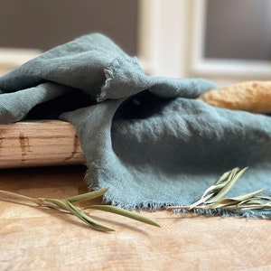 Puede incluir: Un pa&ntilde;o de lino azul verdoso drapeado sobre una cesta de pan de madera con una hogaza de pan dentro. Una ramita de vegetaci&oacute;n est&aacute; en primer plano.