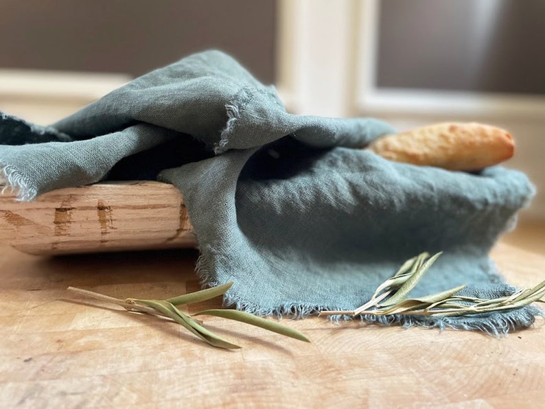 Puede incluir: Un pa&ntilde;o de lino azul verdoso drapeado sobre una cesta de pan de madera con una hogaza de pan dentro. Una ramita de vegetaci&oacute;n est&aacute; en primer plano.