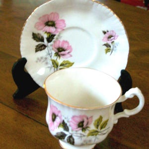 May include: A vintage teacup and saucer set. The white porcelain cup and saucer feature a pink floral design with green leaves and gold trim. The cup has a curved handle and sits on a small foot. The saucer is round.