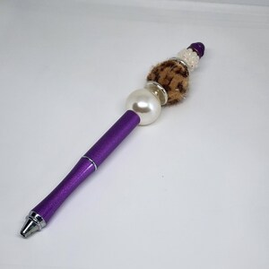 May include: A purple pen with a decorative beaded top. The pen features a large white pearl bead, a brown and tan faux fur accent, and a cluster of white beads. The pen's barrel is a metallic purple.