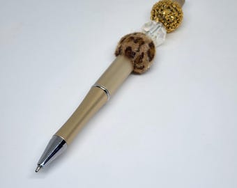 May include: A gold-colored pen with a silver tip. The pen features a decorative design with a leopard print accent, clear beads, and a gold beaded sphere at the top. The pen is designed for writing and is suitable for various occasions.