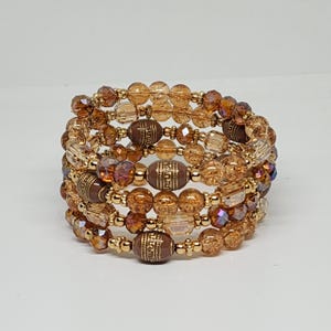 Könnte beinhalten: Ein mehrreihiges Wickelarmband bestehend aus facettierten bernsteinfarbenen Glasperlen, kleinen goldenen Metallzwischenperlen und größeren braunen ovalen Perlen mit detaillierten goldenen Gravurmustern in kreisförmiger Anordnung.