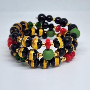 May include: A multi-strand bracelet featuring black and gold striped beads, interspersed with red, green, and black beads. The bracelet is accented with gold-colored spacers and has a coiled design.
