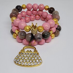 May include: Three stacked beaded bracelets. The bracelets feature pink, gold, and brown beads. A gold crown and a purse charm with clear rhinestones are also included. The bracelets are on a white surface.