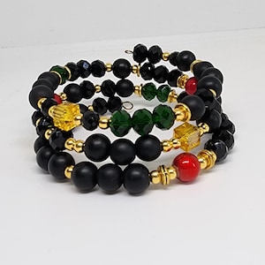 May include: A stack of spiral bracelets featuring black matte and faceted beads, accented with gold-colored metal beads and colorful gems in green, red, and yellow. The bracelets are displayed against a white background.