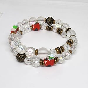 May include: A multi-strand bracelet featuring clear faceted beads, smooth frosted white beads, and antique bronze-toned metal spacers. The design includes small red apple-shaped charms with green leaves and accents of dark textured metal spheres.
