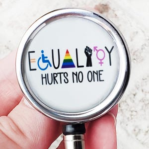 May include: A circular, silver badge reel with a white face. The face displays the word "EQUALITY" with a rainbow-coloured "A" and symbols for disability, LGBTQ+ rights, and gender equality. The text "HURTS NO ONE" is below.