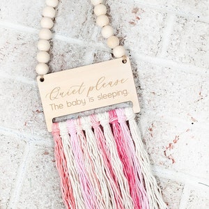 Op de afbeelding: Een houten bord met de tekst "Quiet please The baby is sleeping." hangt aan een houten kralenketting met roze en witte garenkwastjes.