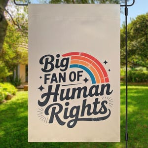 May include: A beige garden flag with the text "Big Fan of Human Rights" in a retro font, with a rainbow graphic. The flag is hanging on a black metal pole in a grassy outdoor setting.