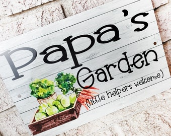 May include: A white wooden sign with black text that reads "Papa's Garden (little helpers welcome)". The sign has a watercolor image of a basket of green apples, carrots, and leafy greens.