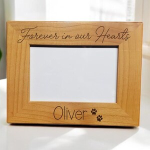 May include: A light wood picture frame with the inscription "Forever in our Hearts" and the name "Oliver" with two paw prints below.