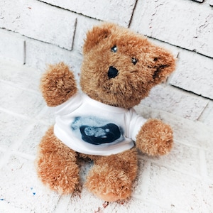 Könnte beinhalten: Ein brauner Teddybär, der ein weißes T-Shirt mit einem schwarz-weißen Ultraschallbild trägt.