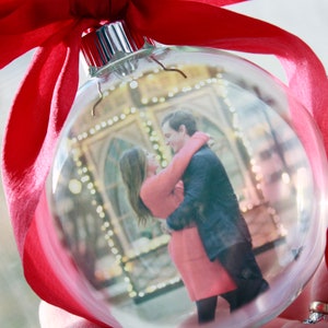 May include: A clear glass Christmas ornament with a photo of a couple inside. The ornament is decorated with a red ribbon.