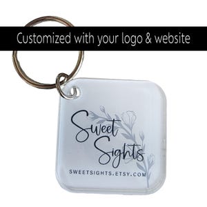 May include: Clear square keychain with a silver keyring. The keychain features the text "Sweet Sights" in black script with a floral design in grey. The website address, sweetsights.etsy.com, is printed below. The top of the image reads "Customized with your logo & website."