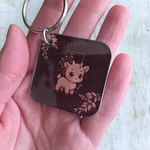 May include: Square keychain featuring a cartoon goat illustration in shades of brown and beige. The goat is inside a rectangular frame with floral accents. The keychain has a silver metal ring for attaching keys.