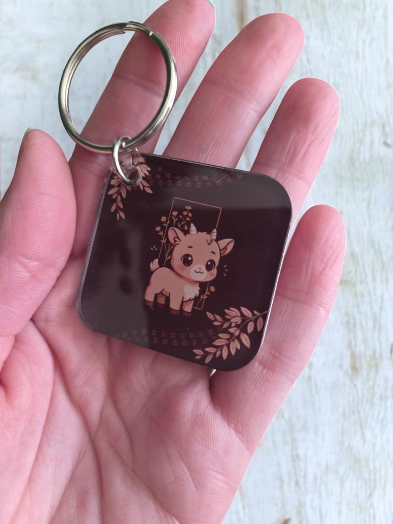 May include: Square keychain featuring a cartoon goat illustration in shades of brown and beige. The goat is inside a rectangular frame with floral accents. The keychain has a silver metal ring for attaching keys.