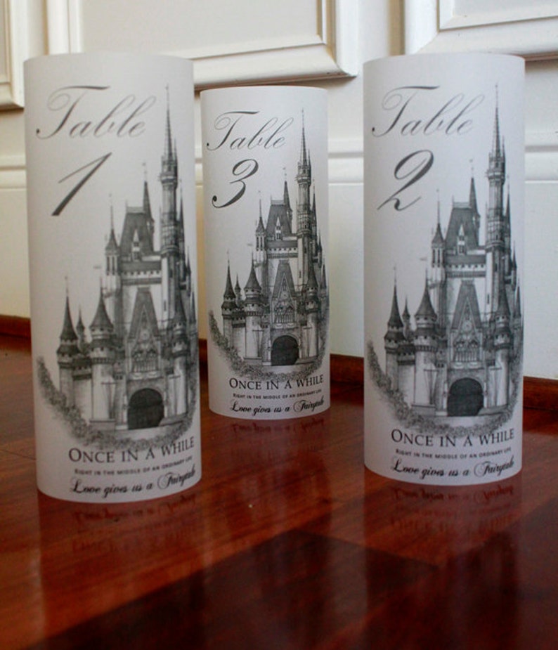 May include: Three white cylinder table number holders with a black and white illustration of Cinderella's castle. The numbers 1, 2, and 3 are written in black script on the front of each holder. The text "Once in a while, right in the middle of an ordinary life, love gives us a fairytale" is printed on the bottom of each holder.