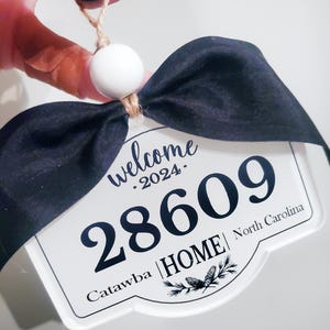 May include: White sign with black text and a black bow. The sign says "welcome . 2024. 28609 Catawba | HOME | North Carolina".