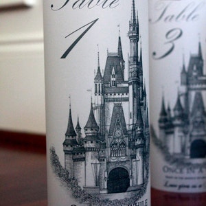 May include: A white cylinder with a black and white illustration of Cinderella's castle. The text "Table 1" is printed in black at the top of the cylinder. The text "Once in a while, right in the middle of an ordinary life, love gives us a fairytale" is printed in black at the bottom of the cylinder.