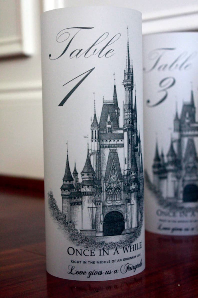 May include: A white cylinder with a black and white illustration of Cinderella's castle. The text "Table 1" is printed in black at the top of the cylinder. The text "Once in a while, right in the middle of an ordinary life, love gives us a fairytale" is printed in black at the bottom of the cylinder.