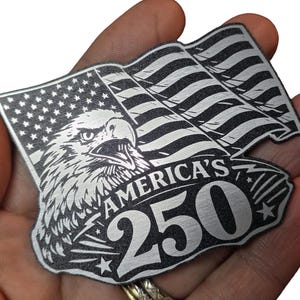 Op de afbeelding: Een zilverkleurig metalen embleem met een Amerikaans vlagontwerp met een adelaar en de tekst "AMERICA'S 250". De vlag wappert en de adelaar kijkt naar voren. Het embleem wordt in een hand gehouden.
