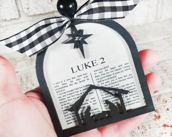 May include: Black and white checkered ribbon tied around a wooden Christmas ornament with a cutout of the nativity scene and the text "LUKE 2" and a portion of the Bible verse from Luke 2:6-14.