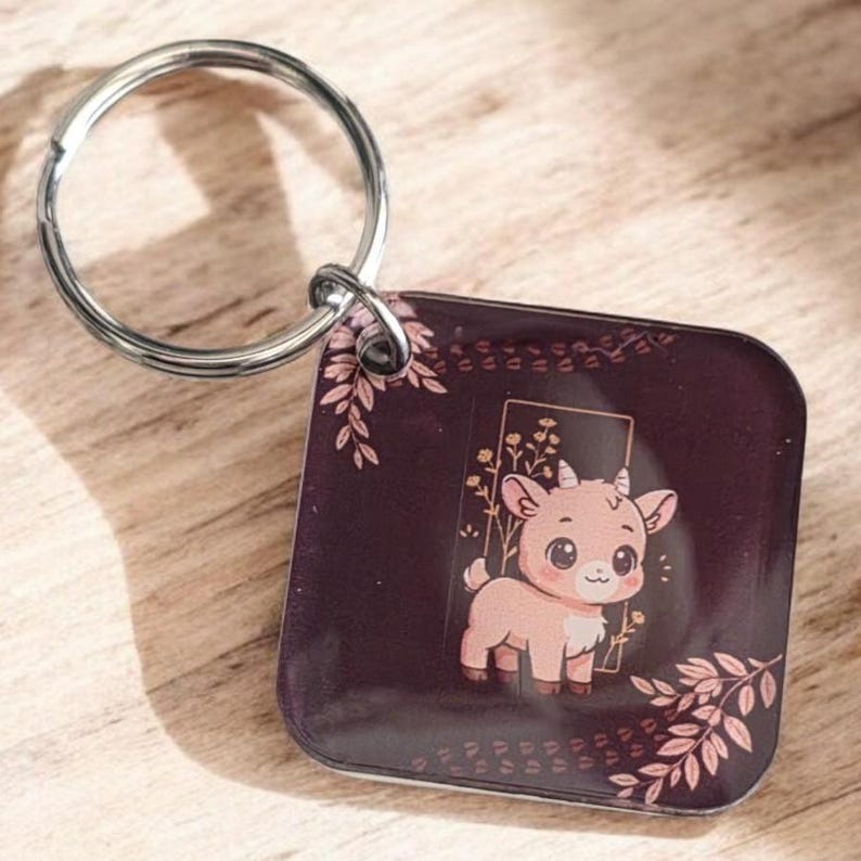 May include: A square keychain with a silver ring features a cartoon goat illustration. The goat is light brown with large eyes, standing in a gold-framed rectangle. The background is dark purple with pink floral accents.