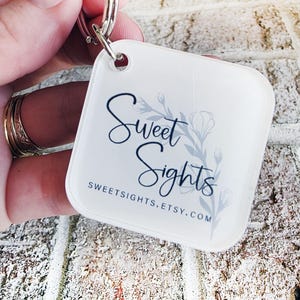 May include: A white, square keyring with rounded corners. The keyring displays the text "Sweet Sights" in a script font, with a floral design and the website address sweetsights.etsy.com.