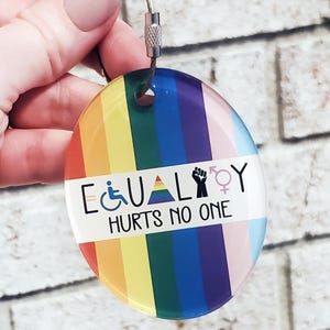May include: A round glass keychain with a rainbow pride flag design. The text "EQUALITY HURTS NO ONE" is printed on a white band across the middle of the keychain. The keychain also features symbols for disability, transgender, and gender equality.