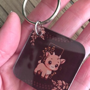 May include: A square keychain with a brown background and a cartoon goat illustration. The goat is light brown with large eyes and small horns. The keychain has a silver metal ring.