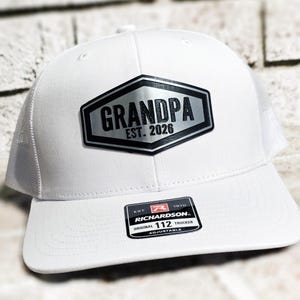Puede incluir: Gorra de camionero blanca con parte trasera de malla y visera curva. La parte delantera presenta un parche hexagonal plateado y negro que dice "GRANDPA EST. 2026". La gorra tiene una correa ajustable.