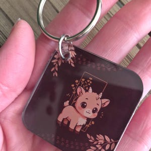 May include: A square keychain with a brown background and a cartoon goat illustration. The goat is light brown with large eyes and small horns. The keychain has a silver metal ring.
