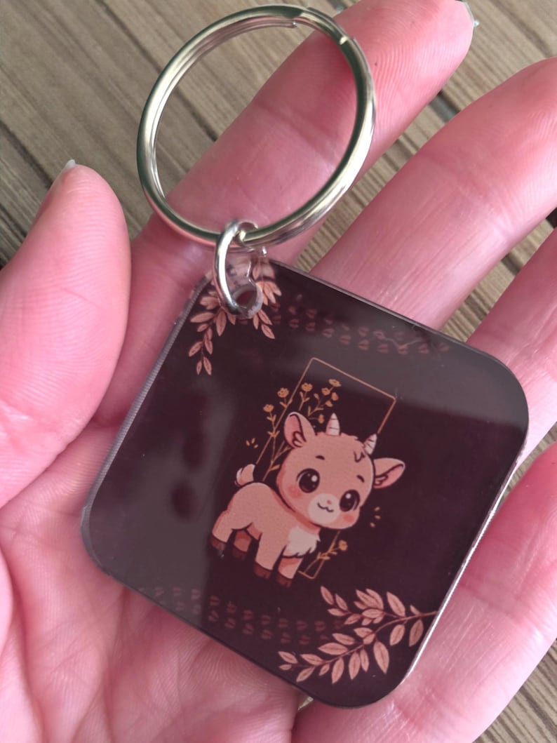May include: A square keychain with a brown background and a cartoon goat illustration. The goat is light brown with large eyes and small horns. The keychain has a silver metal ring.