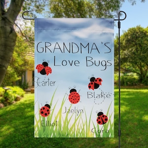 May include: A garden flag with a blue sky background and green grass. The text "Grandma's Love Bugs" is printed in black. Five red ladybugs with black spots are printed on the flag, each with a name underneath: Carter, Jaelyn, Elliot, Blake, and Cora.