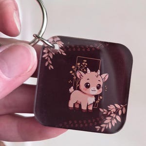 May include: A square keychain with a cartoon goat illustration. The goat is light brown with large eyes and small horns, set against a dark brown background with floral accents. A silver keyring is attached.