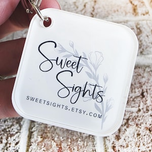 May include: A white, square keyring with rounded corners. The keyring features the text "Sweet Sights" in a script font, with a floral design and the website address SWEETSIGHTS.ETSY.COM. A silver keyring is attached to the top.