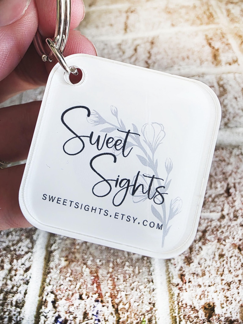 May include: A white, square keyring with rounded corners. The keyring features the text "Sweet Sights" in a script font, with a floral design and the website address SWEETSIGHTS.ETSY.COM. A silver keyring is attached to the top.