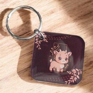 May include: A square keychain with a silver ring features a cartoon goat illustration. The goat is light brown with a white chest and is set against a dark burgundy background with floral accents. The keychain is on a wooden surface.