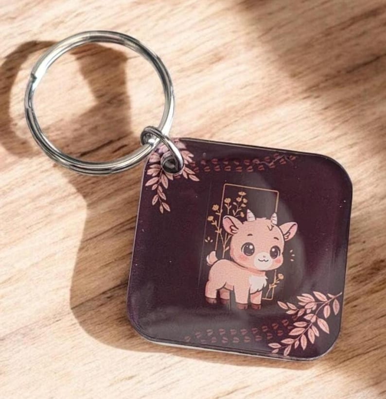May include: A square keychain with a silver ring features a cartoon goat illustration. The goat is light brown with a white chest and is set against a dark burgundy background with floral accents. The keychain is on a wooden surface.