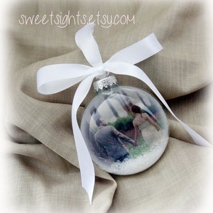 May include: A clear glass ornament with a white ribbon and a photo of a couple inside. The ornament is filled with white glitter.